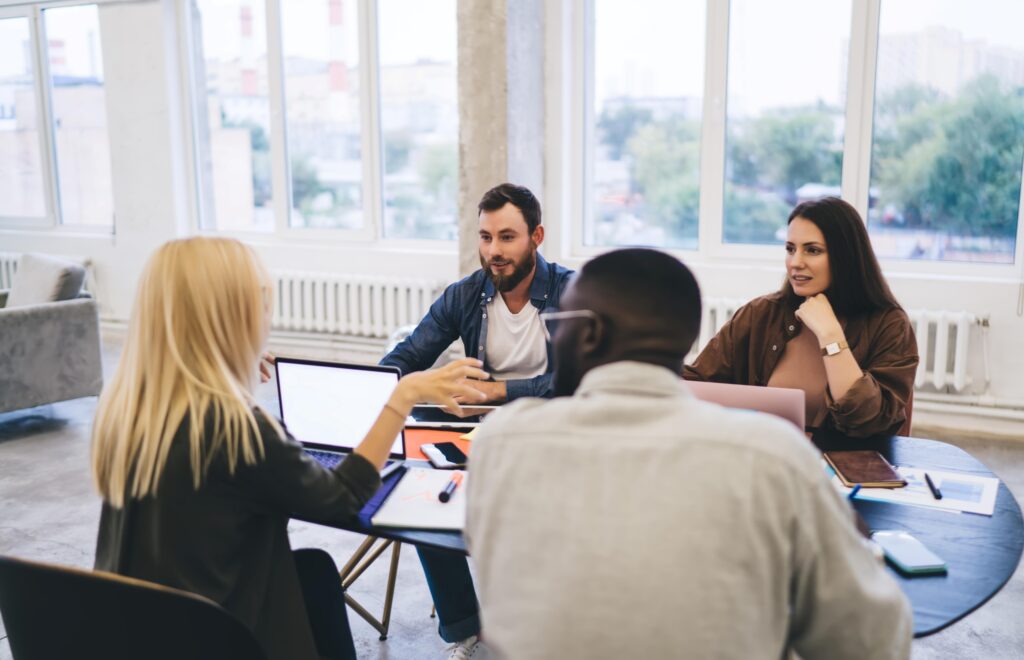 A group of 4 people sitting around a table with laptops and having a meeting.