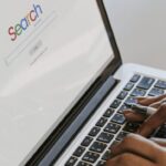 A picture of someone typing on a laptop with a visual of a search engines home screen on the lap top.
