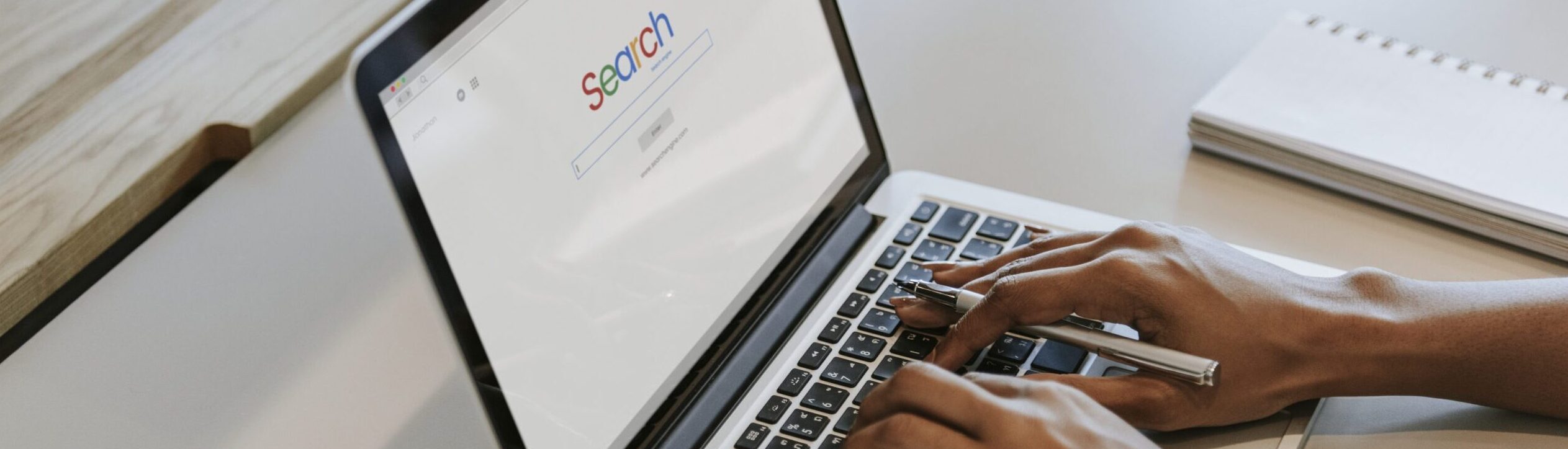 A picture of someone typing on a laptop with a visual of a search engines home screen on the lap top.